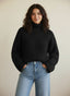 Woman in a black turtleneck sweater and light blue jeans standing against a neutral background.