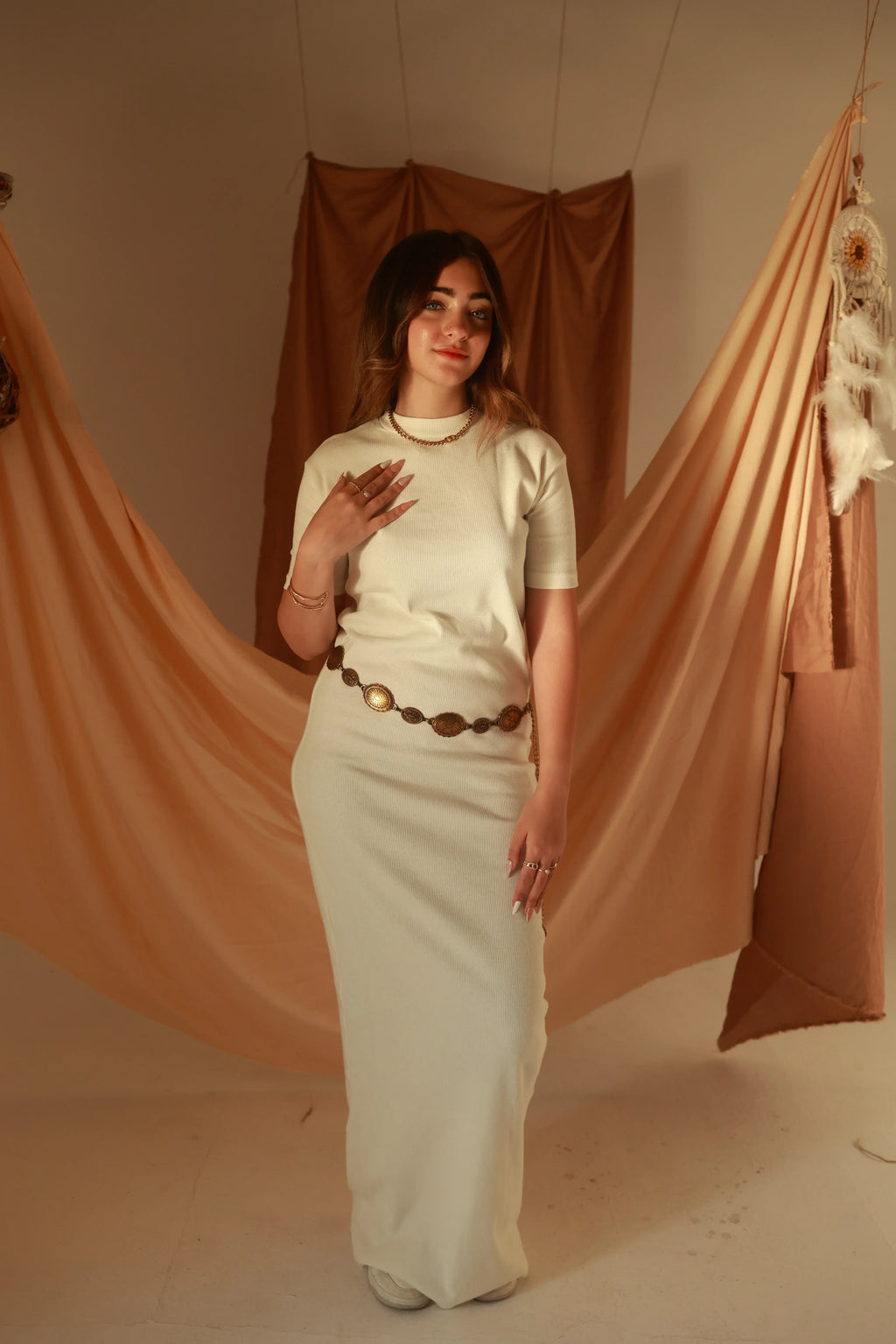 Woman in cream knit maxi dress with gold jewelry and brown belt, neutral draped backdrop