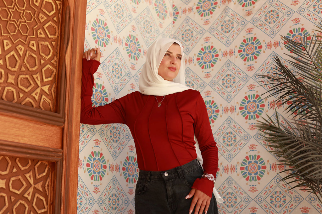 Woman in a white hijab and red long-sleeve top posing by a carved wooden door, floral patterned wall in background