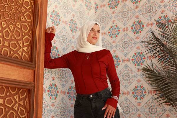Woman in a white hijab and red long-sleeve top posing by a carved wooden door, floral patterned wall in background